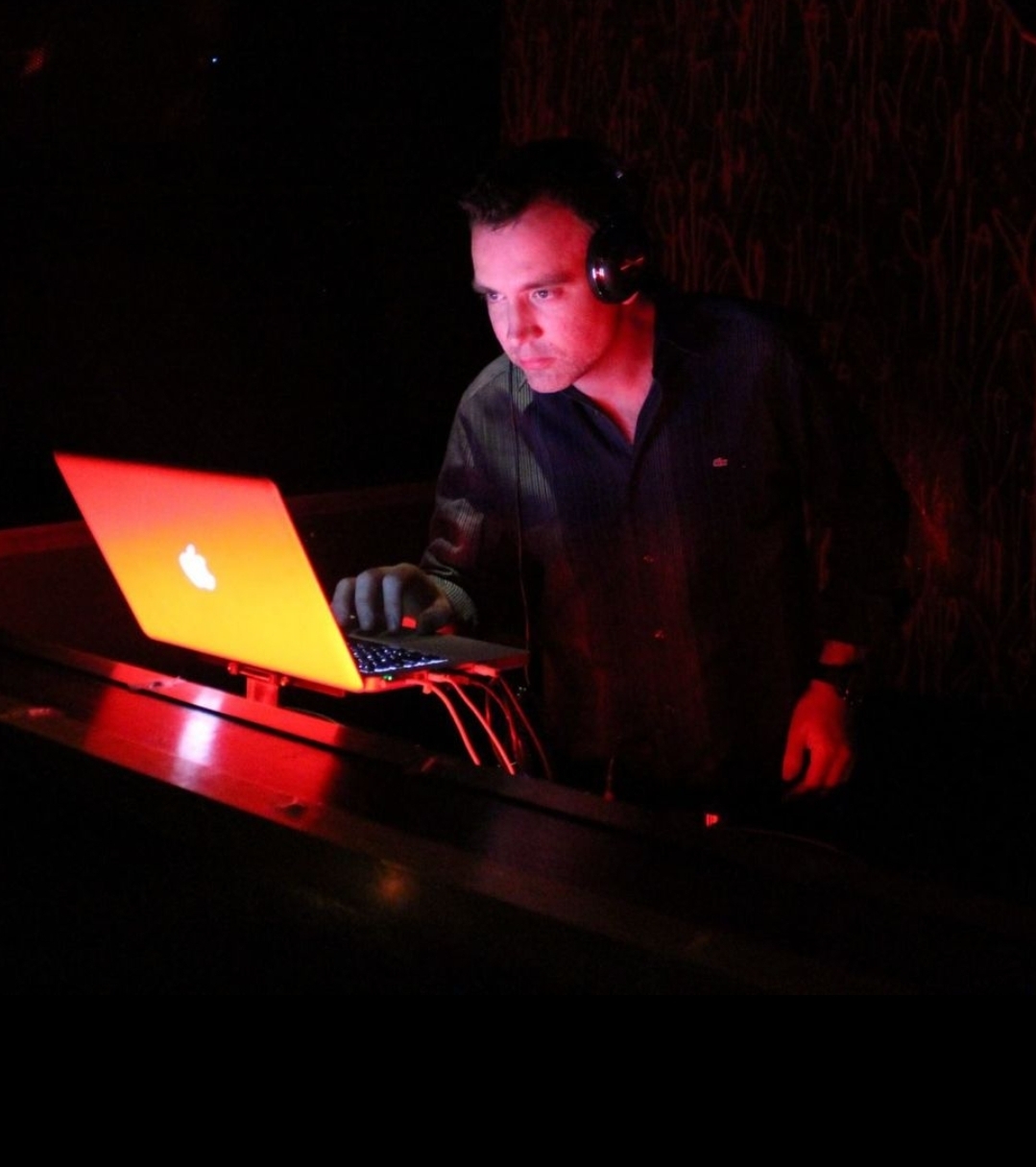 Ashley Heather DJing with headphones while operating a laptop in a dimly red-lit venue.
