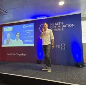 Ashley Heather speaking into a microphone on stage during a presentation at the Health Optimisation Summit.